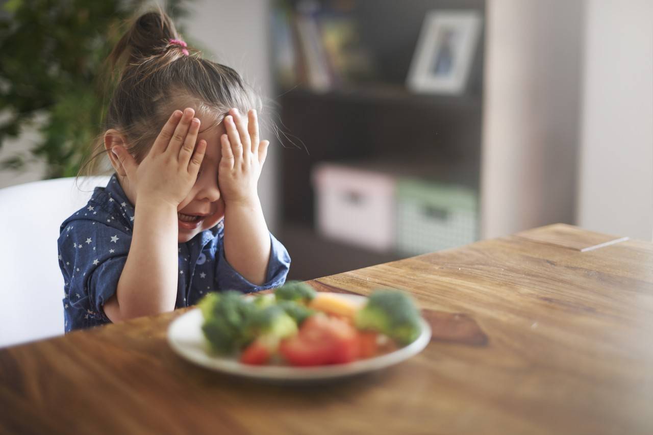 menina de dois anos sentada na mesa se recusando a comer um prato com brócolis e outros vegetais