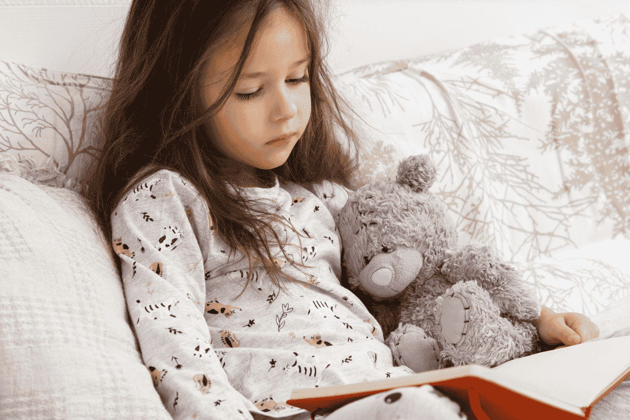 menina de pijama lendo livro com ursinho de pelúcia